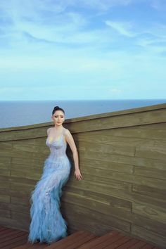 a woman in a blue dress leaning against a wooden wall next to the ocean with her hands on her hips