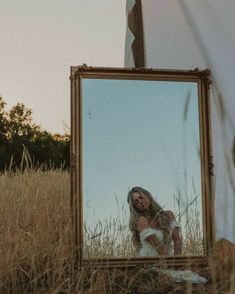 a woman standing in front of a mirror on top of a grass covered field with trees