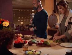 a man and woman are preparing food on a kitchen table while two other people stand in the background