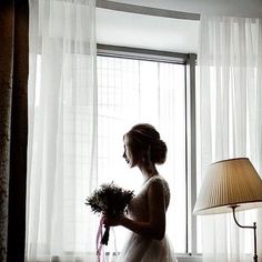 a woman standing in front of a window next to a lamp and holding a bouquet