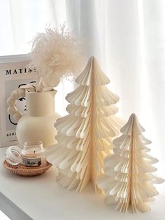 three paper christmas trees on a table next to a book and magazine holder with feathers in it