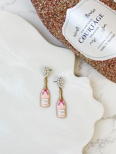 a pair of pink and gold earrings sitting on top of a white marble table next to a bottle of champagne