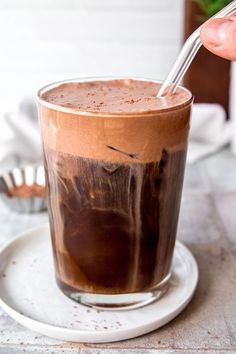 a hand holding a spoon over a chocolate drink in a glass on a saucer