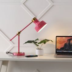 a desk with a laptop, lamp and plant on it in front of a white wall