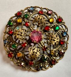 This is an Edwardian era brooch of Czech origin. It features ornate layered brass filigree openwork and is decorated with prong set multi-coloured Czech glass gems in red, blue, green, yellow.   The central red stone measures 0.8cm across and the brooch measures 6cm in diameter (2.36 inches). It fastens with a c-clasp. The brooch is in very good condition, with normal signs of wear and age. All stones are present. Era 1910s Condition Excellent Antique Multicolor Brooch For Gifts, Antique Multicolor Brooches As Gifts, Vintage Multicolor Multi-stone Brooch, Vintage Multi-stone Gold Brooches, Vintage Gold Multi-stone Brooches, Vintage Multicolor Brooches, Antique Multicolor Brooch Jewelry, Victorian Style Multicolor Brooches As Gift, Vintage Multicolor Collectible Brooches