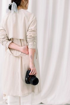 a woman holding a camera in her right hand and wearing a beige trench coat with sheer sleeves