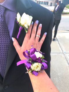 two purple and white wrist corsages with flowers on the arm, one being held by a woman's hand