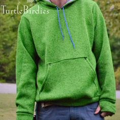 a man wearing a green hoodie standing in front of some trees and grass with his hands on his hips