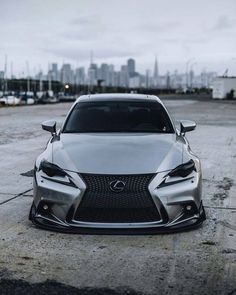 the front end of a silver car parked in a parking lot with a city skyline behind it