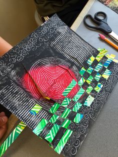 a person holding up a piece of art with scissors on the table next to it