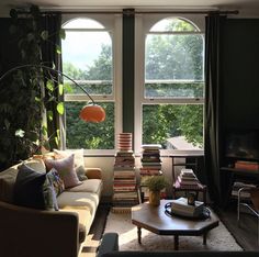 a living room filled with furniture next to two large windows covered in green curtains and plants