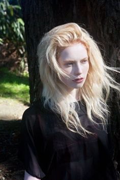 a woman with blonde hair standing in front of a tree and looking at the camera