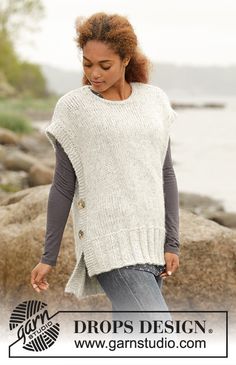 a woman wearing a white sweater and jeans standing on rocks next to the water with her hands in her pockets