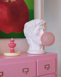 a pink dresser topped with a white busturine next to a red apple painting