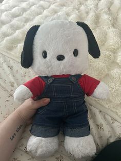 a white stuffed dog wearing overalls on top of a bed