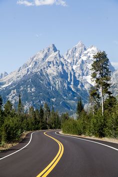 an empty road in the mountains with snow on the mountain tops behind it and trees