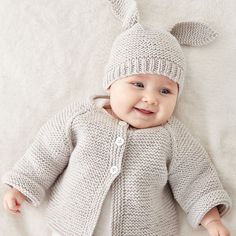 a smiling baby wearing a knitted bunny hat and cardigan laying on a white blanket