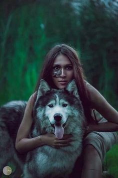 a woman with makeup on her face sitting next to a wolf in the grass and holding it's head