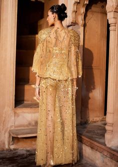 A bold three piece dress in sheer fabric, a glamorous embellished gold bralet paired with sheer trousers with splatter embroidery of sitara/patra and a matching sheer jacket with power shoulder highlighted with gold embellishments Festive Chic Sets With Cape Sleeves, Organza Party Sets With Cape Sleeves, Glamorous Evening Sharara With Dupatta, Evening Floor-length Palazzo Set With Mirror Work, Evening Palazzo Set With Cape Sleeves And Dupatta, Hand Embellished Evening Sets For Eid, Festive Evening Set With Cape Sleeves, Festive Evening Palazzo Set With Cape Sleeves, Elegant Embellished Palazzo Set For Party