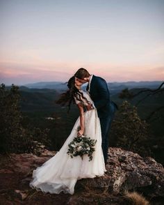 a bride and groom kissing on top of a mountain at sunset with the sun setting behind them