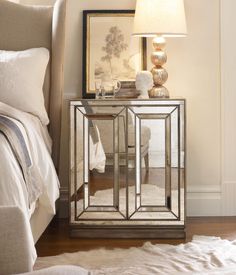 a mirrored side table with a lamp on it next to a white bed and pillows