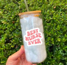 a person holding up a glass with the words best nurse ever on it in front of bushes