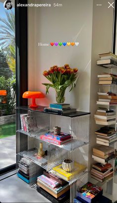 a book shelf filled with lots of books next to a large window and a vase full of flowers