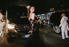 a woman in a long black dress standing next to a car on the street at night