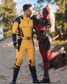 two people dressed as deadpool and spider - man standing in the sand with trees in the background