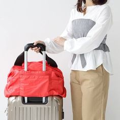 A woman standing with a grey suitcase with a large red bag through the handle she holds. Practical Large Capacity Travel Accessories For On-the-go, Foldable Nylon Travel Bag For Everyday Use, Functional Foldable Travel Bag For Daily Use, Packable Nylon Bags For Weekend Trips, Foldable Travel Bag For Everyday Use, Practical Foldable Nylon Travel Bag, Packable Nylon Duffle Bag For Travel, Multifunctional Large Capacity Travel Bag, Foldable Rectangular Shoulder Bag For Outdoor Activities