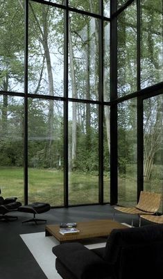 a living room filled with black furniture and large glass windows that look out onto the woods