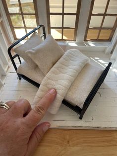 a hand pointing at a miniature bed made out of blankets on a wooden floor with windows in the background