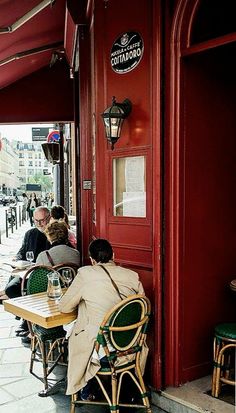 people sitting at tables in front of a red door on a sidewalk with green chairs