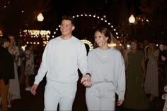 a man and woman are holding hands while walking through the street with lights in the background