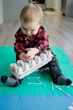 a toddler sitting on the floor playing with an egg carton and knitting needles