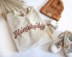 Homebody's unite Live your best lounge life in this cozy crew neck sweatshirt. It is sure to keep you feeling all warm and fuzzy all season long. Beige Letter Print Sweatshirt For Loungewear, Cream Letter Print Sweatshirt For Loungewear, Trendy Cream Sweatshirt For Loungewear, Trendy Cream Sweatshirt, Cute Sweatshirt Designs, Homebody Shirt, Beige Sweatshirt, Western Tee, Summer Graphic Tee