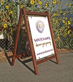 a wooden sign sitting on the side of a road in front of a chain link fence