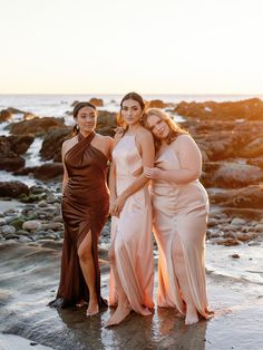 three women in dresses standing on the beach at sunset with their arms around each other