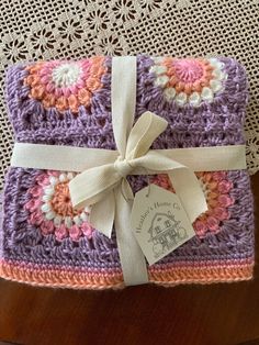 a purple and pink crocheted blanket with a white ribbon on the end, sitting on a table