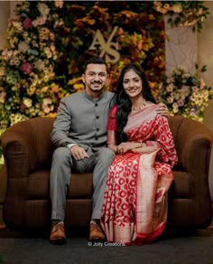 a man and woman sitting on a couch in front of a floral wall with flowers