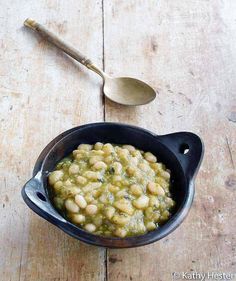 a bowl filled with beans and greens next to a spoon on a wooden table top