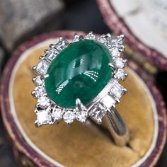 This magnificent ring is centered with one (1) oval cabochon cut natural emerald weighing 5.18 carats and set into a four-prong setting. The emerald is bordered with eight (8), prong set, square step cut diamonds and twelve (12), prong set, round brilliant cut diamonds. The ring measures 23.1mm at the top, rises 12.2mm above the finger, tapering to 3.8mm wide and 1.9mm thick at the base of the shank. This ring is currently a size 7.75. Oval Platinum Emerald Ring With 17 Jewels, Platinum Oval Emerald Ring With 17 Jewels, Green Oval Emerald Ring In Platinum, Luxury Oval Emerald Gemstones, Luxury Oval Cabochon Emerald Ring, Luxury Oval Emerald Birthstone Ring, Luxury Emerald Ring With Oval Cabochon And 17 Jewels, Luxury Oval Cabochon Emerald Ring With 17 Jewels, Luxury Emerald Ring With Oval Cabochon