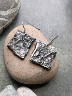 VORA GRISOS Earrings Reticulated metal Hand chiselled Patinated silver Solid silver ear hooks Delivered in its red and gold pouch Maintains like classic silver with a dry and soft cloth, or with silverware if necessary. I used bronze plates that I had in my workshop and I sawed small square shapes. I then heated them with a torch to a certain temperature, so that the metal melted but not completely and I managed my flame to give them this textured appearance. This is called cross-linking. I then reworked them, using small goldsmith chisels of abstract shapes. The metal was then completely stripped using chemical processes. The silvering was made by electrolysis in my workshop in Nice. Their shape is unique, and the two loops are different in appearance which gives them a more aesthetic. Yo Jewellery Earrings, Earrings Geometric, Earrings Metal, Bronze Metal, Metal Hand, Recycled Metal, Ear Hook, Metal Earrings, Chic Woman