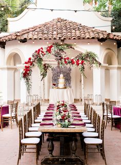 a table set up for a wedding with red flowers and greenery on the top