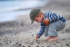 a little boy playing on the beach with pebbles in his hands and wearing a hat