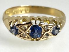 Sapphire & Diamond Ring This stunning antique ring from the George V era features a beautiful oval-shaped sapphire stone with a round cut sapphire on either side, in a yellow gold setting. The ring is further adorned with four diamonds, making it a perfect choice for any special occasion such as Mother's Day, Christmas, Graduation, Anniversary, Birthday, or Valentine's Day. Trilogy rings traditionally represent the past the present and the future. The ring is hallmarked dating it to 1918 and it Antique Blue Oval Diamond Ring, Vintage Oval Sapphire Ring Hallmarked, Antique Oval Sapphire Diamond Ring, Vintage Oval Sapphire Ring With Hallmark, Antique Oval Sapphire Ring With Rose Cut Diamonds, Victorian Sapphire Ring In Yellow Gold, Victorian Oval Sapphire Ring, Victorian Style Yellow Gold Sapphire Ring, Victorian Style Oval Sapphire Ring