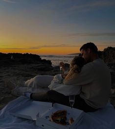 a man and woman sitting on the beach at sunset with pizza in front of them