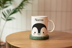 a coffee mug with a penguin on it sitting on top of a wooden table next to a potted plant