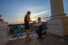 a man kneeling down next to a woman on top of a balcony with a sunset in the background