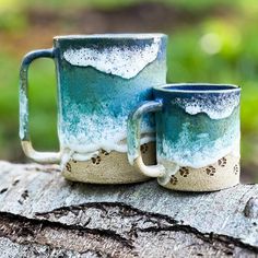 two coffee mugs sitting on top of a tree stump in the woods, one has blue and white glaze